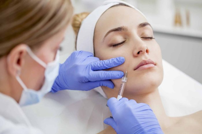 A woman lies with her eyes closed while a professional wearing blue gloves and a face mask injects a substance into her cheek with a syringe. The woman has a white headband on and appears relaxed during the cosmetic procedure.