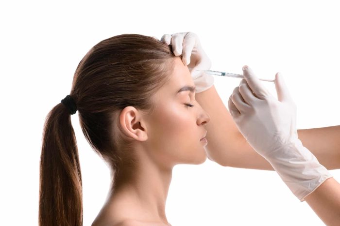 A person with long brown hair in a ponytail receives a cosmetic injection on their forehead. They have their eyes closed, and a medical professional wearing white gloves administers the injection. The background is white and uncluttered.