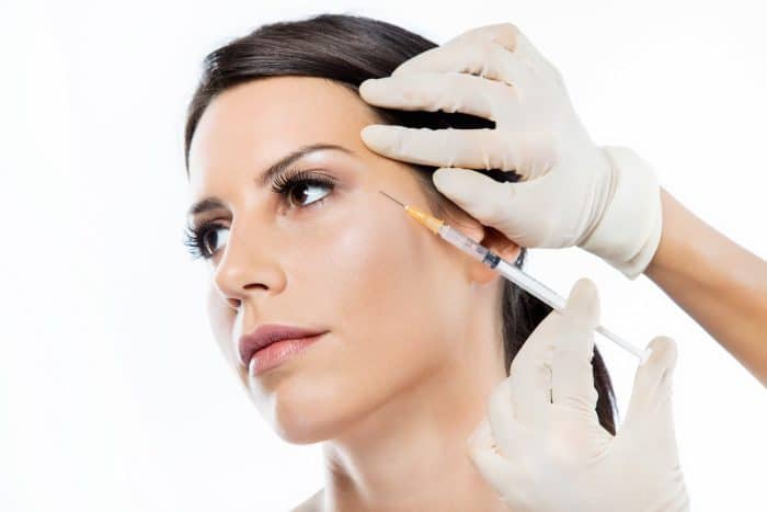 A close-up of a woman receiving a cosmetic injection in the temple area of her face, potentially botox for TMJ treatment. She has dark hair pulled back and is looking to the side. The person administering the injection is wearing white gloves and holding a syringe filled with a clear substance.