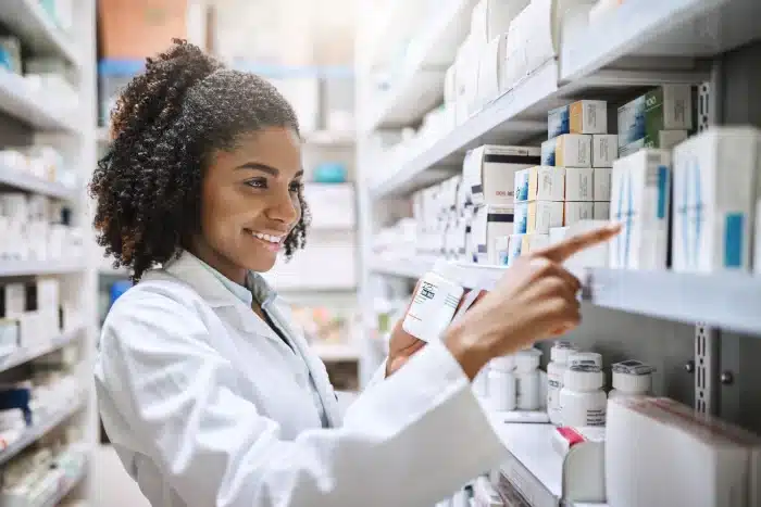 A pharmacist wearing a white lab coat stands in front of medicine shelves, smiling while holding a medication bottle in one hand and reaching for another box with the other hand. The shelves are stocked with various pharmaceutical products, including options for Xeomin vs Botox treatments.