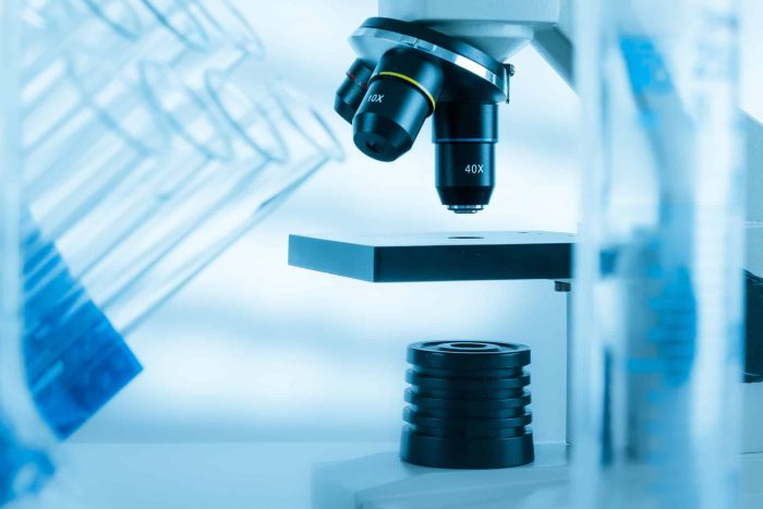 A close-up of a microscope set at 40x magnification is seen on a laboratory bench. There are several test tubes blurred in the foreground and background, giving the image a scientific and clinical atmosphere. The lighting is cool and blue-tinted, hinting at research into artificial ovaries.