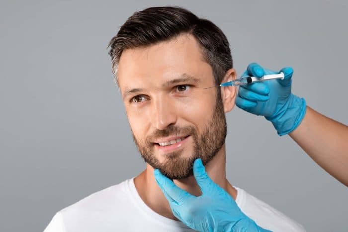 A man with short hair and a beard smiles as another person in blue gloves administers the best under eye filler with a syringe near his eye. The background is a plain, light gray.