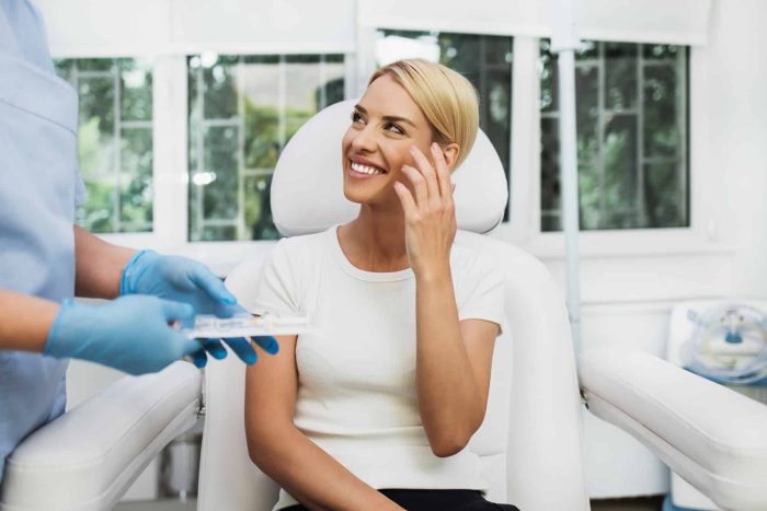 A woman with short blonde hair sits in a medical chair, smiling and touching her face. She is looking at a healthcare professional wearing gloves who is holding a syringe. The background shows a window with greenery outside.