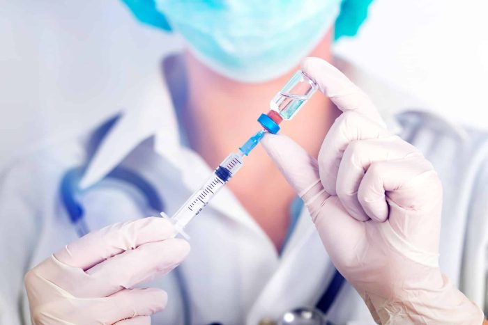 A medical professional wearing a mask and gloves holds a syringe and vial, preparing to administer a vaccine. The background is blurred, focusing attention on the syringe and vial.