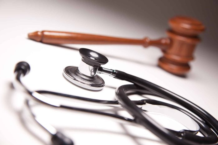 A stethoscope lies on a white surface with its chestpiece in focus, while a wooden gavel rests in the background. The image symbolizes the intersection of healthcare and legal matters.