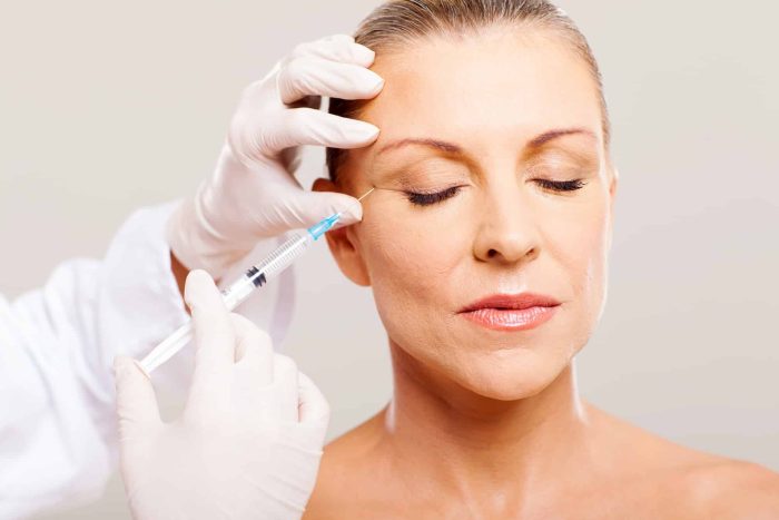 A healthcare professional wearing white gloves administers an injection to a woman's face using a syringe. The woman, with her eyes closed and hair pulled back, appears calm and relaxed. The background is plain and light-colored.