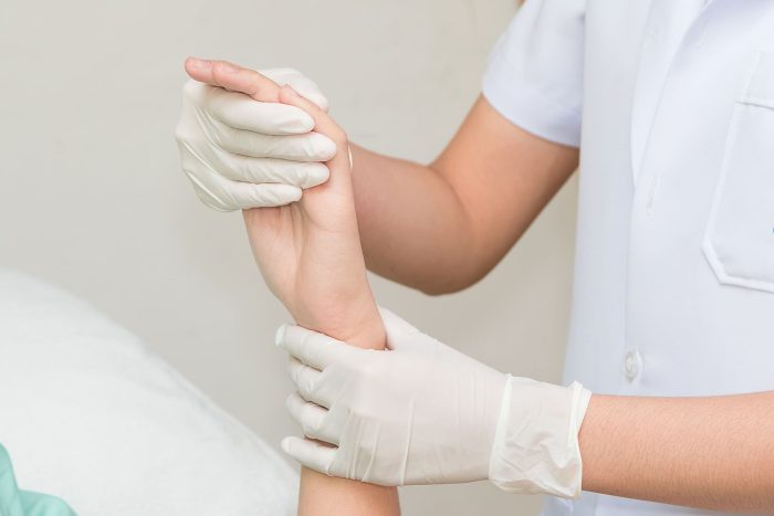 A healthcare professional wearing a white uniform and gloves is holding the wrist of a patient, gently lifting the hand. The patient's arm is extended, and the setting appears to be a medical examination or treatment environment.