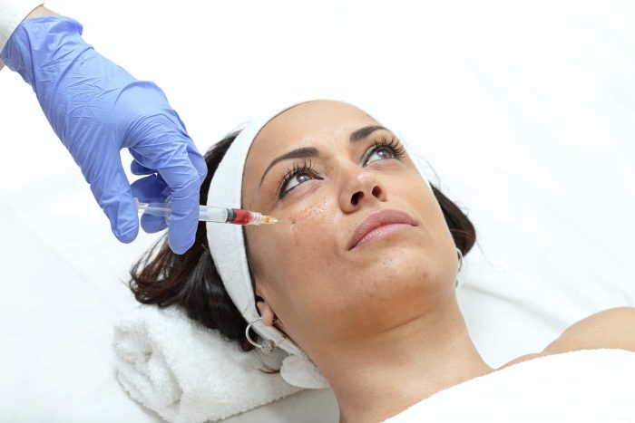 A woman lies on a treatment table with a white headband and towel. A gloved hand is holding a syringe near her face, likely preparing for a cosmetic or medical injection. The setting appears to be a professional medical or spa environment.