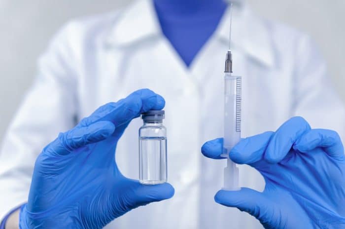 A person wearing a white lab coat and blue gloves holds a vial of clear liquid in one hand and a syringe in the other, perhaps preparing for a Restylane vs Botox treatment. The focus is on the hands and the medical items, with the person's face blurred in the background.