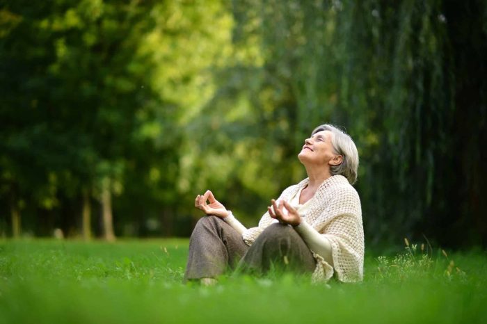 An elderly person sits cross-legged on the grass in a park, eyes closed, head tilted upwards, and smiling peacefully. They are wearing a light-colored shawl. Sunlight filters through the trees in the background, creating a serene atmosphere.