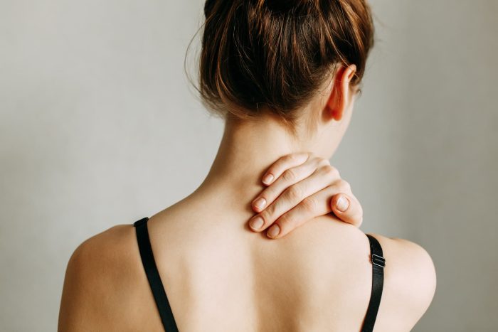 A person with light brown hair is shown from behind, with their hair in a bun. They are wearing a black spaghetti strap top and have one hand placed on the back of their neck, appearing to massage or soothe it. The background is plain and light-colored.