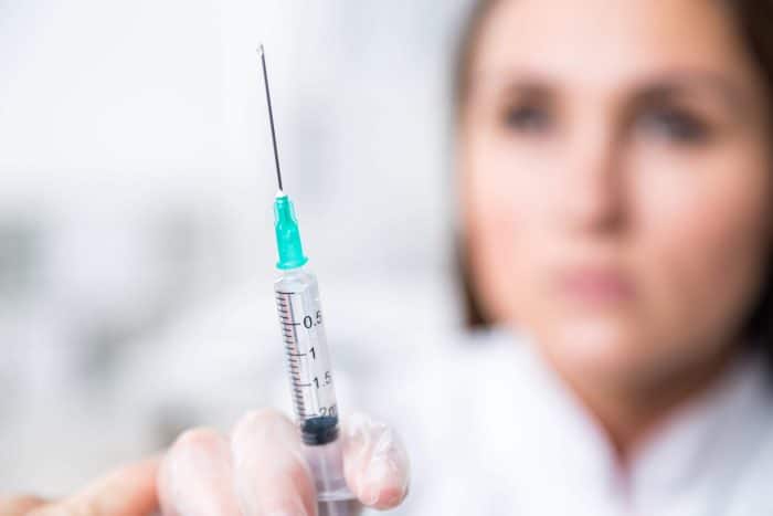 A person in medical attire, partially blurred, holds a syringe with a green cap and a measurement scale. The syringe is in sharp focus, highlighting the needle and clear fluid inside. The background is soft and out of focus, subtly hinting at the choice between Juvederm vs Botox for treatments.