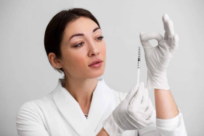 A woman wearing a white lab coat and gloves holds up a syringe, examining it closely against a white background. With a focused expression and her dark hair pulled back, she appears to be demonstrating how to recognize authentic filler products.