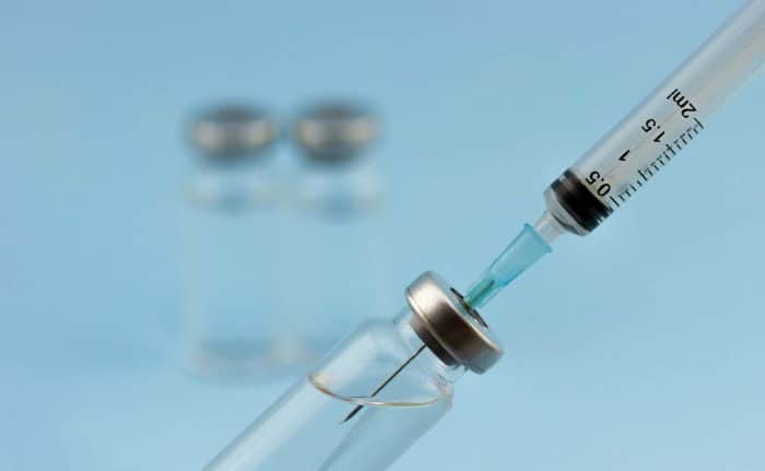 A syringe drawing liquid from a vial with a metal cap. Two additional blurred vials are in the background against a light blue surface.