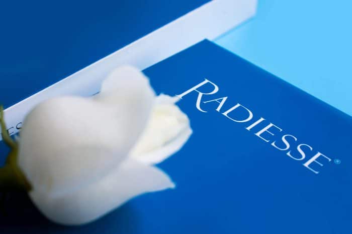 A close-up of a blue Radiesse branded box with a defocused white rose in the foreground. The image focuses on the box's logo, subtly highlighting its radiesse filler packaging against a light blue background, with slightly blurred edges to add depth.