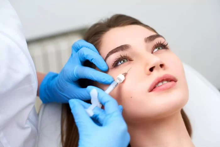 A person with brown hair is lying back while a medical professional wearing blue gloves administers a Restylane tear trough injection near the person's eye. The environment appears to be a clinical setting.