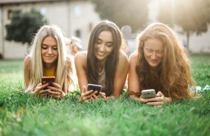 Three young women lying on grass in a park, smiling and looking at their smartphones. They appear relaxed and happy, with sunlight creating a warm atmosphere in the background. Trees and a building can be seen in the distance as they enjoy capturing their self image.