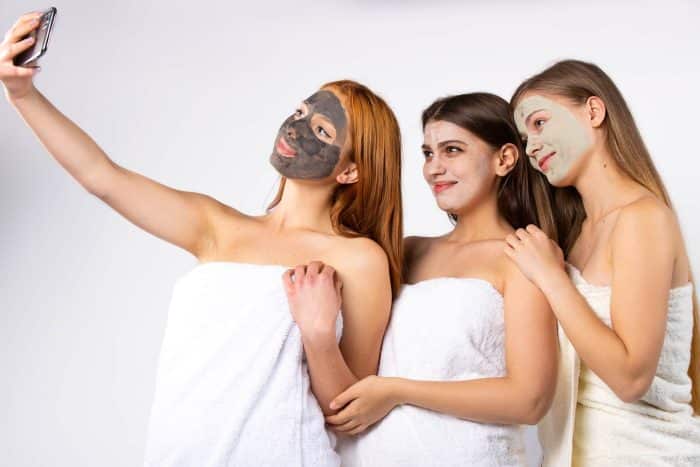 Three women wearing towels and facial masks stand together taking a selfie, showcasing facial aesthetics for millennials. One woman sports a black mask, the second a white one, and the third a green mask. They are smiling against a plain white background.