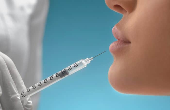 Close-up of a medical professional holding a syringe near the lips of a person. The person's face is depicted in profile with attention focused on the lower part of the face and the syringe, reflecting current lip filler trends. The background is a soft blue gradient.