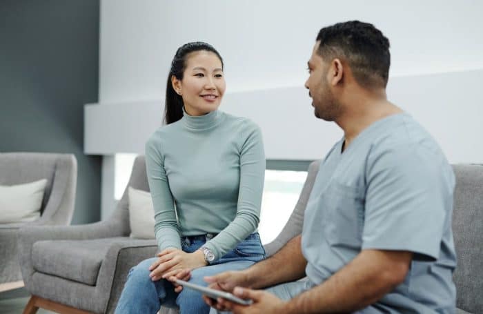 A woman in a light blue sweater and jeans sits and smiles while speaking with a man in medical scrubs who is holding a tablet. They are seated on gray chairs in a white and gray waiting area, discussing her upcoming aesthetic consultation.