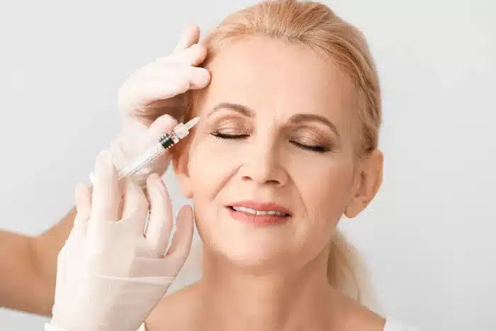 A person with closed eyes receiving a cosmetic injection in their forehead from a gloved professional. The person has blonde hair, light makeup, and appears calm, suggesting a medical or aesthetic treatment setting.