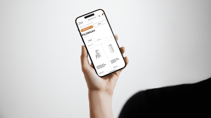 A person is holding a smartphone that displays a website for a skincare brand. The screen shows search filters and two skincare products, one being a bottle and the other a tube, with the Filorga price prominently highlighted. The background is plain white.