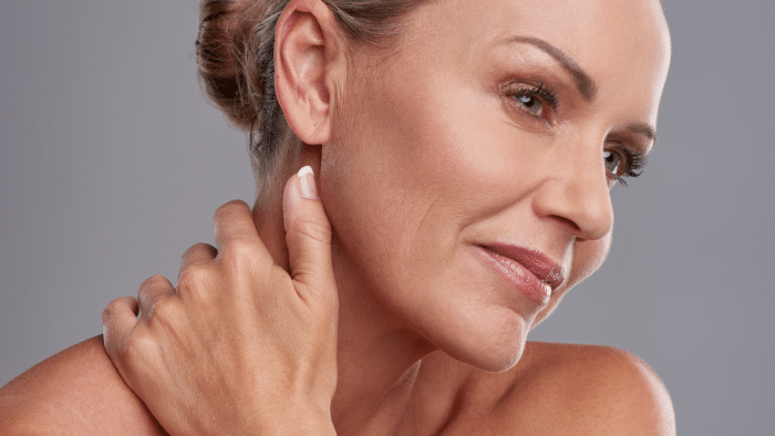 Close-up of a middle-aged person with smooth skin, looking contemplatively to the side. They are touching their neck with one hand and have neatly styled hair pulled back. Wearing neutral makeup, the individual has a serene expression against a gray background, highlighting the results of Filorga top products.