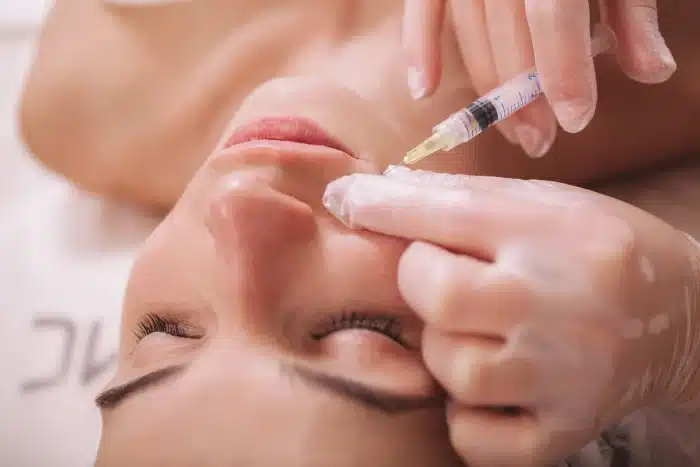 A person is lying down with their eyes closed, receiving an injection in the facial area from a gloved hand. The scene suggests a cosmetic or medical procedure, focusing on the close-up of the face and the syringe.