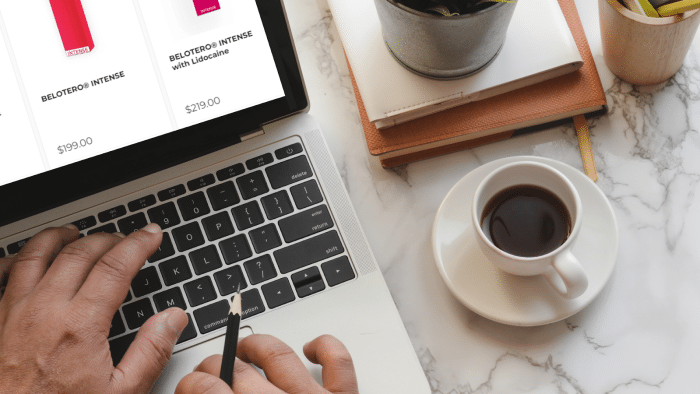 A person types on a laptop displaying a shopping website. Next to the laptop, there is a small white cup of black coffee, a stack of notebooks, a plant in a pot, and other office items—perhaps some research on under eye filler or Belotero products–on the white marble tabletop.