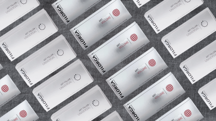 A repeated pattern of FILORGA brand packaging laid out diagonally on a gray carpet. The predominantly white packaging with black and red text reads "Art Filler Universal" and "Art Filler Volume" with circular design elements, subtly hinting at the premium filorga injection price.