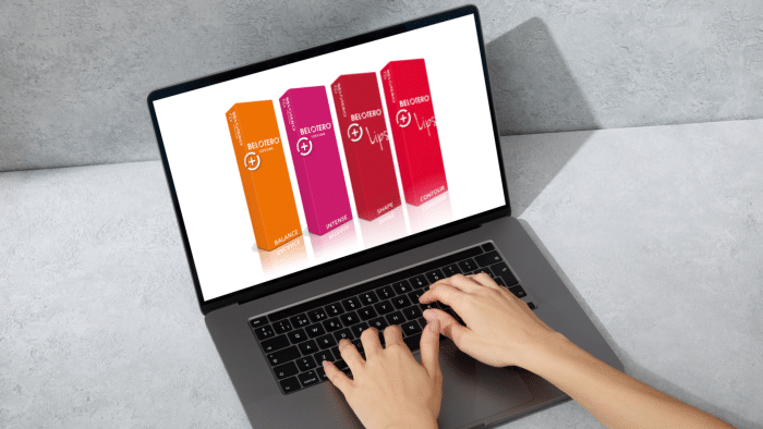 A person is typing on a laptop with a screen displaying four colorful product boxes standing upright. The boxes are orange, red, purple, and red-pink, each with white logos and text. Among them is the sleek packaging for Belotero lip filler, against a textured gray background.