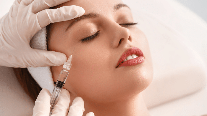 A person lying down with closed eyes receives a cosmetic injection on the cheek from a professional in white gloves. The individual appears relaxed and calm, epitomizing a serene beauty procedure setting. This image perfectly captures the belotero before and after transformation.