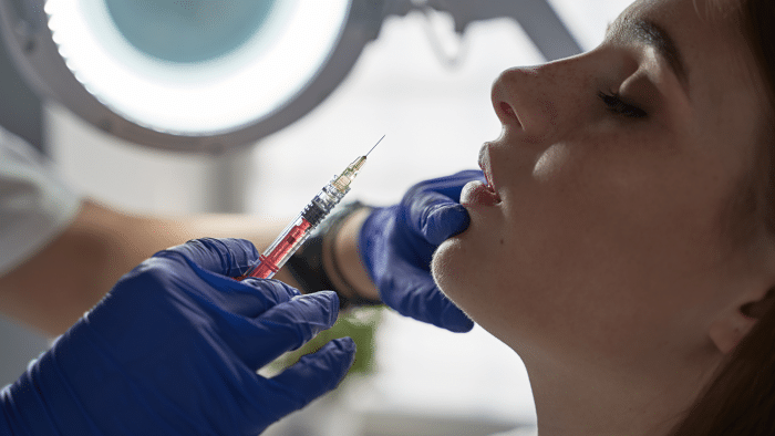 A person with closed eyes receives a facial injection from a professional wearing blue gloves. The professional uses a syringe filled with a red liquid, possibly an anti-aging treatment like Filorga. A bright circular light is visible in the background.