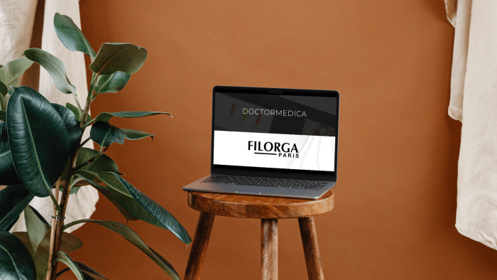 A laptop displaying the Filorga Paris logo is placed on a wooden stool against a warm, brown backdrop. A green plant with large leaves is situated on the left side of the scene, adding a touch of nature to the minimalist setup, subtly prompting viewers to wonder: where can I buy Filorga products?