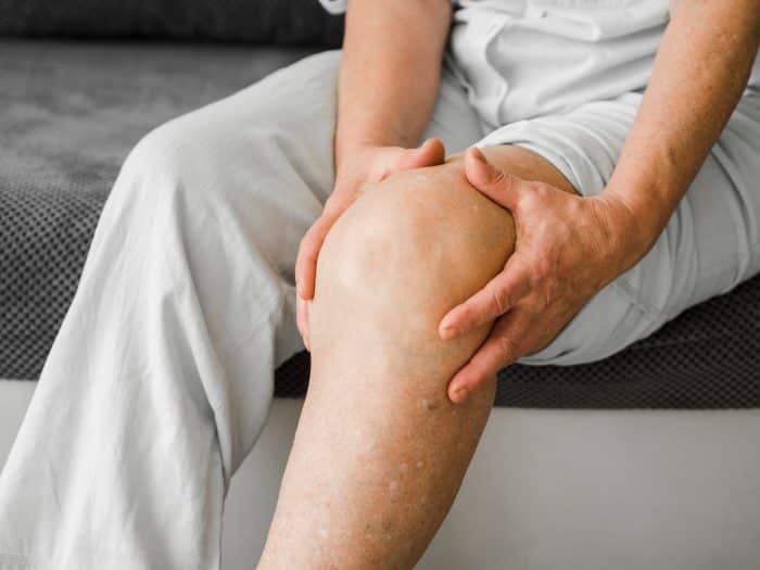 An older individual sits on a couch with both hands placed on their right knee, appearing to hold or massage it. The person, wearing light-colored pants and a shirt, seems to be contemplating the pain relief that Orthovisc injections used for arthritis might offer. The knee is positioned prominently in the center of the image.