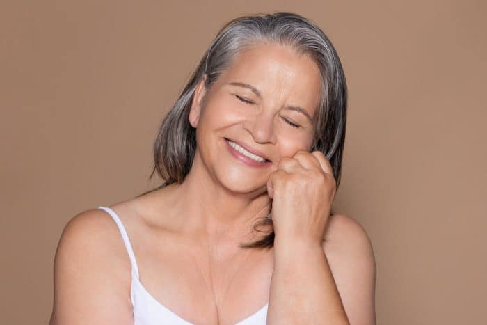 An older woman with gray hair, potentially curious about how quickly botox does its magic, smiles warmly with her eyes closed and her chin resting on her hand. She wears a white tank top, and the background is a solid light brown color.