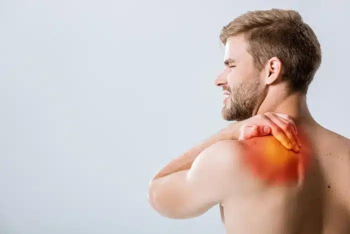 A shirtless man with a beard, seen from behind, is touching his right shoulder with his left hand. The shoulder area shows a red and yellow highlight, indicating pain or inflammation. The man appears to be wincing, suggesting discomfort or pain.