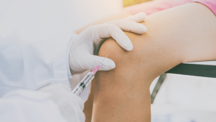 A healthcare professional wearing white gloves administers an injection into a patient's knee, possibly wondering, "Does Orthovisc really work?" The patient is seated with their knee exposed. The background is softly lit.