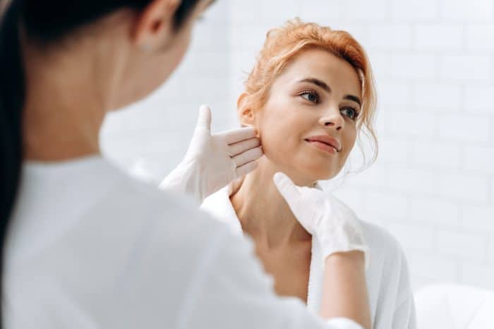 A woman with red hair tied back is having her face and neck examined by a healthcare professional wearing white gloves. They are in a well-lit, clinical environment with white-tiled walls. The woman looks content and relaxed, anticipating the maximum effect from her Botox treatment.