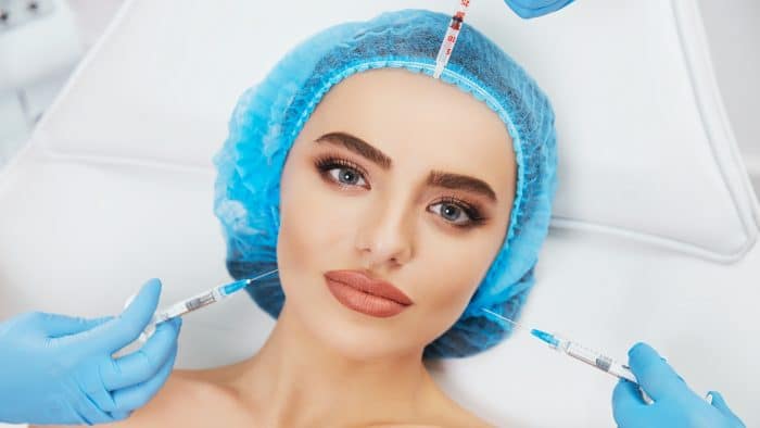 A woman wearing a blue hair cover and lying down is receiving botox injections from gloved hands, aiming for immediate results. She has noticeable makeup with bold eyebrows and lipstick. Three syringe needles are near her face, one on the forehead and two on each side of her cheekbones.