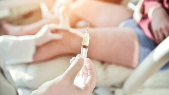 A medical professional in gloves holds a syringe filled with a yellow liquid, preparing for an injection. Another person wearing shorts is partially visible in the background, seated or lying down with legs exposed, possibly indicating what to expect after an Orthovisc injection.