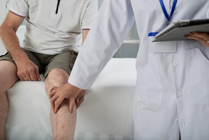 A healthcare professional in a white coat examines a patient's knee. The patient, wearing a light-colored polo shirt and shorts, is seated on an examination table. While touching the patient's leg and holding a tablet, the professional discusses if Orthovisc really works.