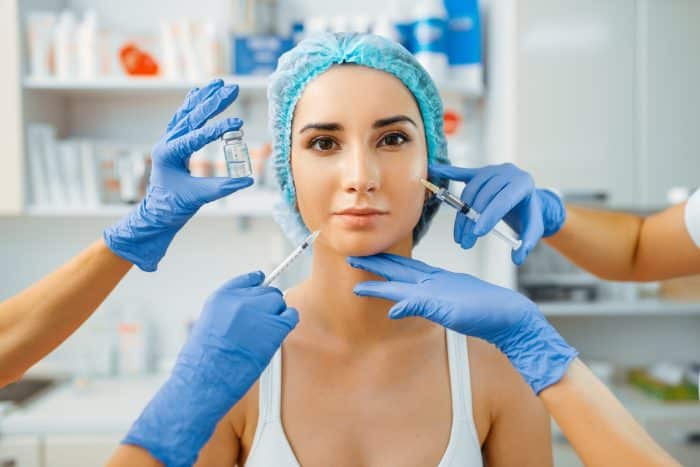 A woman in a medical setting is receiving multiple Botox injections. She wears a blue surgical cap and white tank top, while three gloved hands, each holding a syringe, approach her face and neck. Shelves with various medical supplies illustrate how to prepare for the procedure in the background.