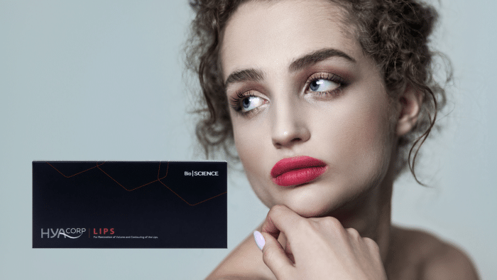 A woman with curly hair and bright red lipstick looks thoughtfully to the side. To her left, there is a black product box labeled "HYACORP LIPS" and "BioSCIENCE," designed for lip augmentation and contouring. She's considering writing a HYACORP LIPS review while admiring its sleek packaging.