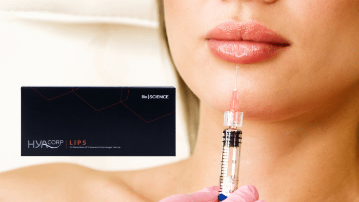 A close-up shot captures a woman's lips as she receives an injection with Hyacorp Lips. A gloved hand holds the syringe near her mouth, while a box labeled "HYACORP LIPS Bio SCIENCE" is visible to the left, indicating the product being used. The background remains blurred for focus.