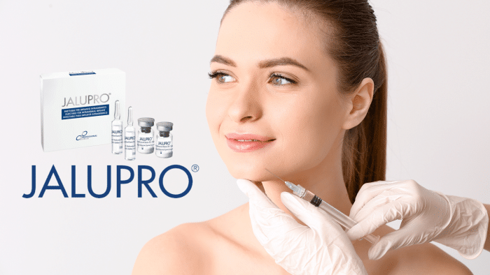 A person receives an injection in the cheek from gloved hands. Beside them is a box labeled "JALUPRO," along with several vials and an ampoule, highlighting the beneficial Jalupro ingredients. The image promotes a skincare treatment product, with a close-up focus on the person’s face and the product packaging.