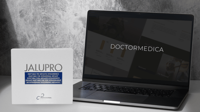 A laptop displaying the DOCTORMEDICA website is open on a desk with a grey textured wall in the background. Next to the laptop, there is a white box labeled "JALUPRO," highlighting its multilingual instructions for those looking to buy JALUPRO online.