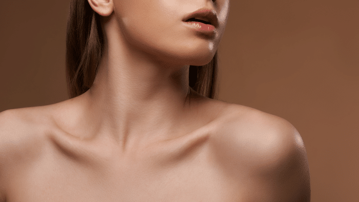 Close-up of a woman’s lower face, neck, and shoulders against a muted brown background. Her smooth skin and slightly parted lips create an alluring focus. The composition emphasizes the contours and lines of the neck and collarbones, highlighting the effects of Jalupro neck treatment.