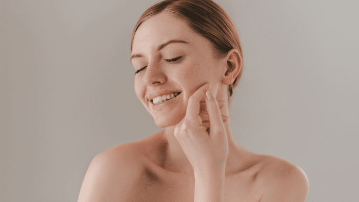 A woman with a serene expression and closed eyes touches her cheek with her hand. She has light skin, light brown hair pulled back, and she appears to be enjoying the impressive Jalupro results of her skincare routine. The background is neutral and simple.