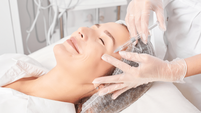 A woman is lying on a medical bed with her eyes closed, smiling, while a healthcare professional wearing gloves administers an injection to her face near the eye area. The woman, possibly experiencing positive jalupro treatment reviews, is wearing a hair cap, and medical equipment is visible in the background.